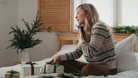 caucasian woman wrapping christmas gift and talking on phone while sitting on bed.