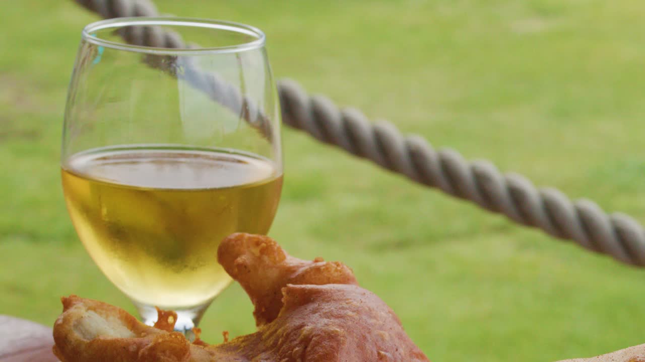 A glass of light beer is poured beside crispy fried fish and chips with salad on a wooden table in a bright outdoor setting