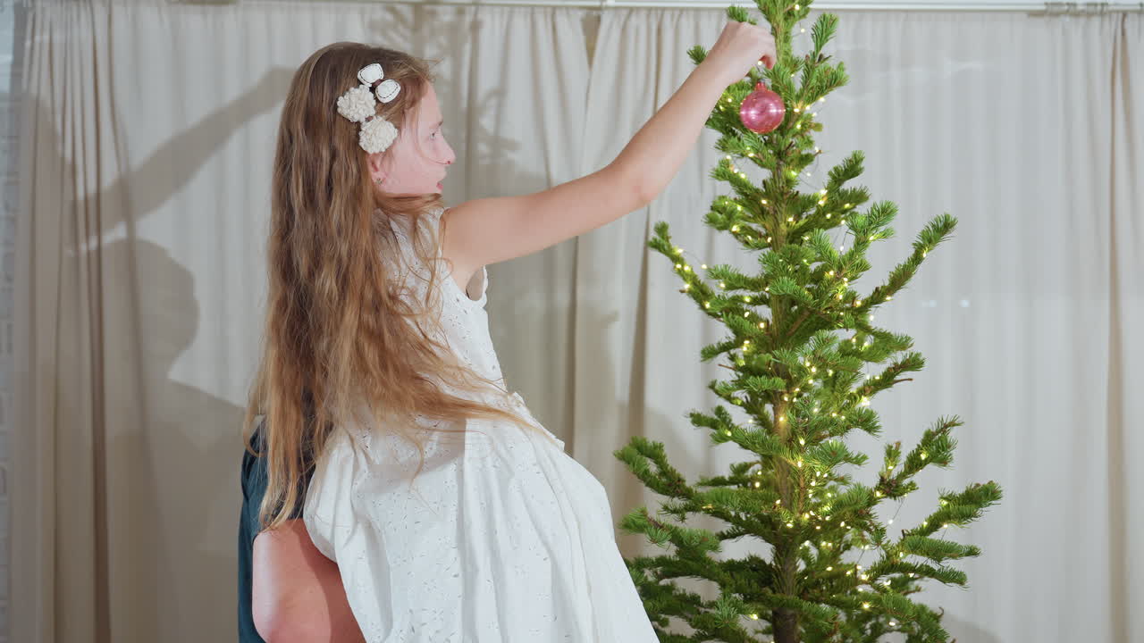 Loving dad carrying daughter in arms helping her place shiny ornament on beautiful christmas tree with twinkling lights during cozy family holiday decorating moment inside warm festive