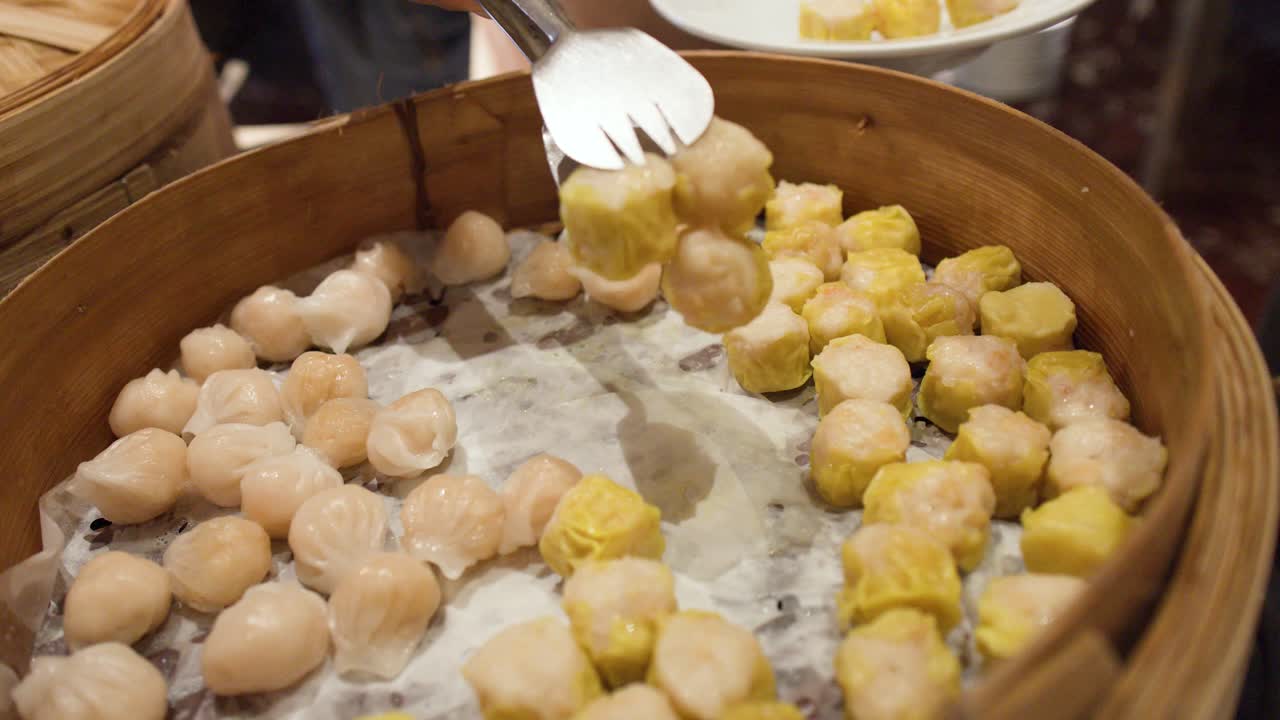 Close-up shot of a person selecting yellow shumai dumplings from a bamboo steamer basket