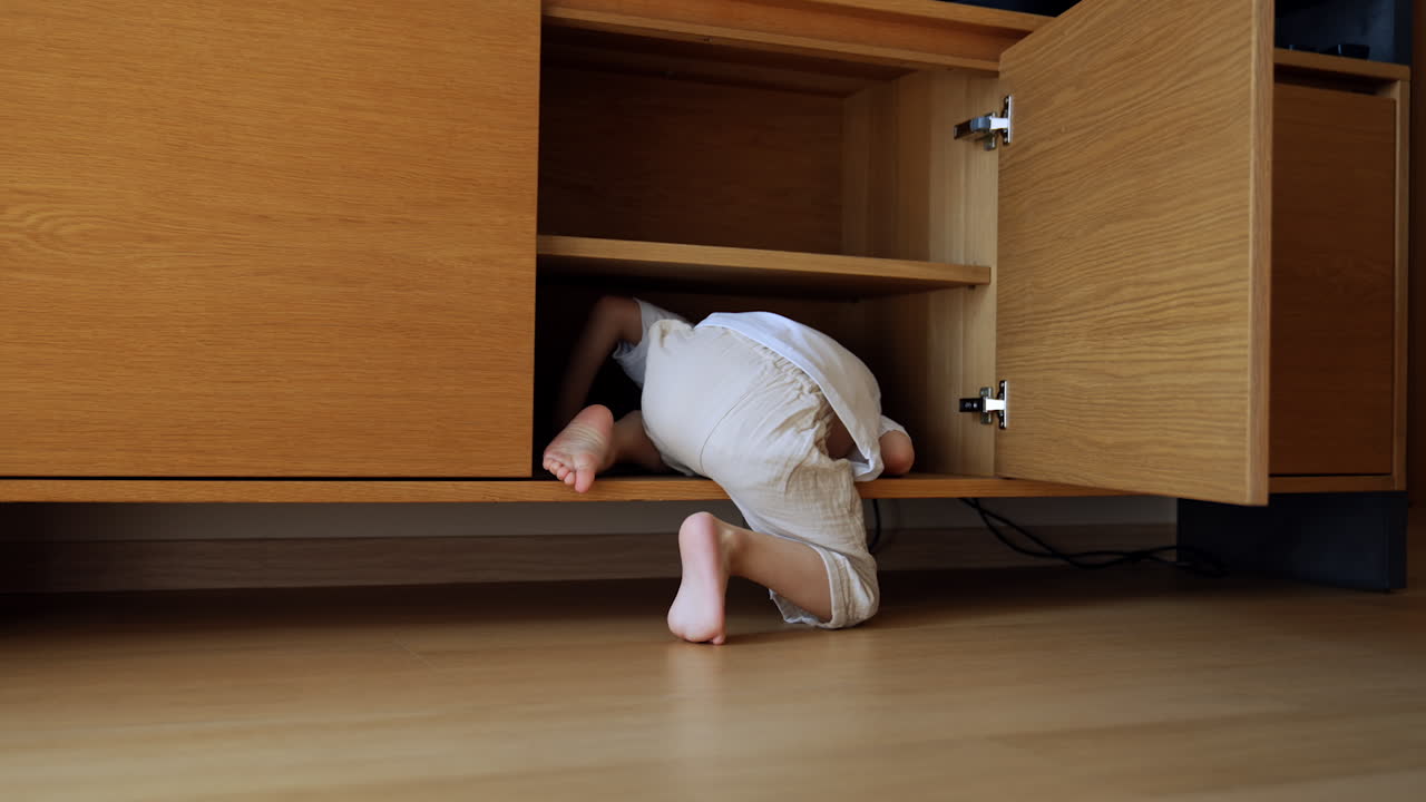 Caucasian toddler wearing white t-shirt and pants hides in the large commode. Active kid plays happily at home