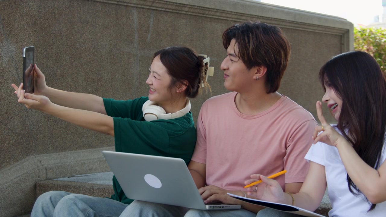 3人の学生がキャンパスの階段に座り、ノートパソコンとタブレットを使って屋外でグループ学習をしています。