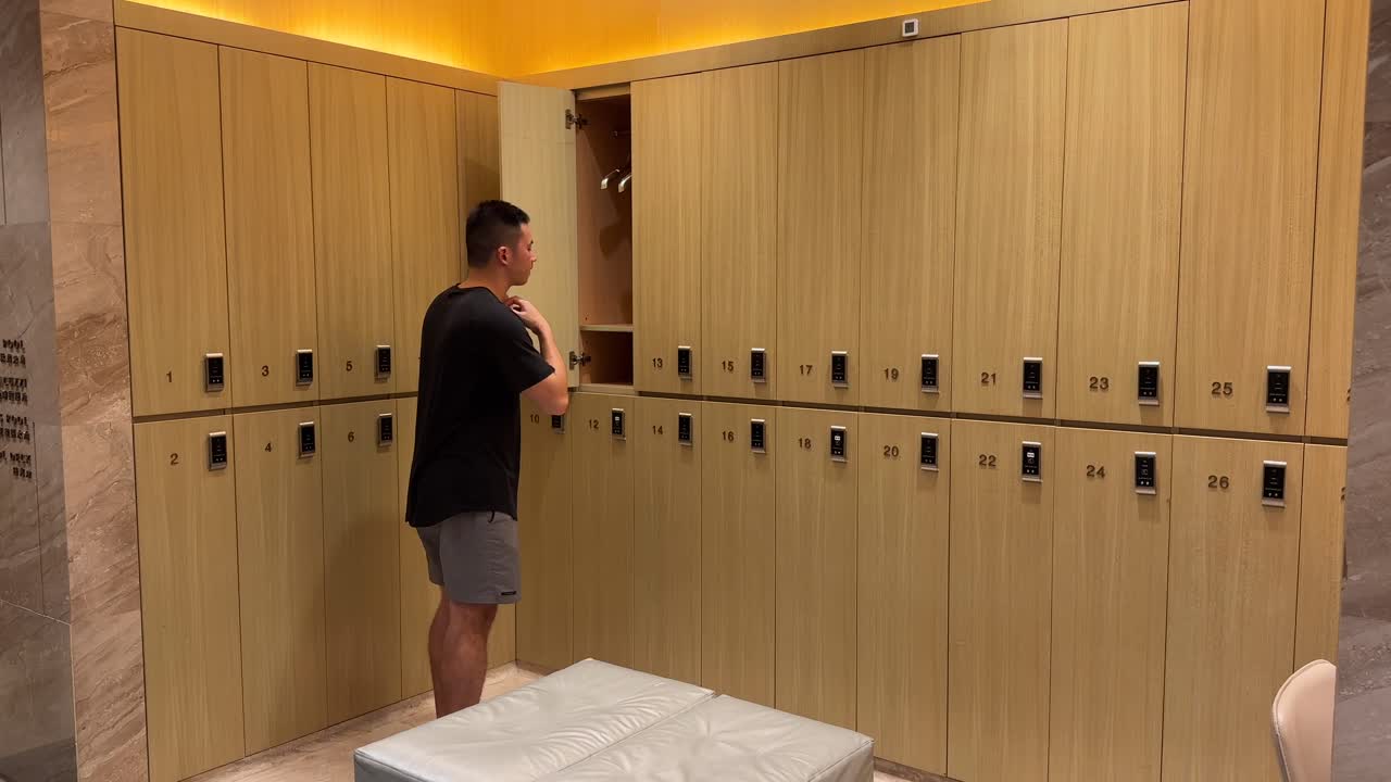 아시안 밀레니얼 남자가 옷을 벗고 셔츠를 고급스러운 옷을 갈아입는 방의 로커에 매달고 있습니다.