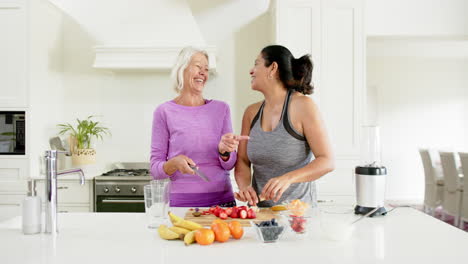 Dos-Mujeres-Mayores-Felices-Y-Diversas-Cortando-Frutas-Y-Riendo-En-Una-Cocina-Soleada,-En-Cámara-Lenta