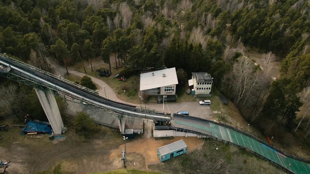 Prospettiva aerea che rivela una vecchia struttura abbandonata di trampolino da sci circondata da una fitta pineta, mostrando l'impianto sportivo in rovina dalla torre di lancio fino alla collina di atterraggio