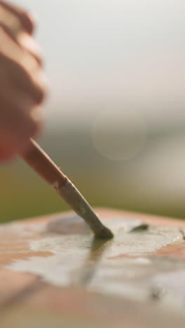 a close-up shot of a woman's hand, with red-painted nails, carefully mixing colors on a wooden palette with a paintbrush under natural sunlight. the scene captures the precise moment of color blending