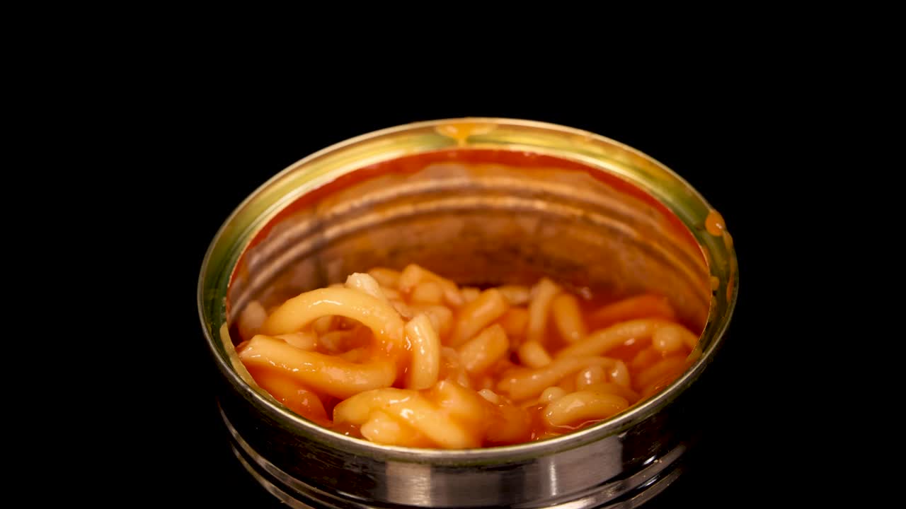 A metal fork lifts saucy canned spaghetti from an open tin, isolated on a black background with bright, even lighting and a static camera