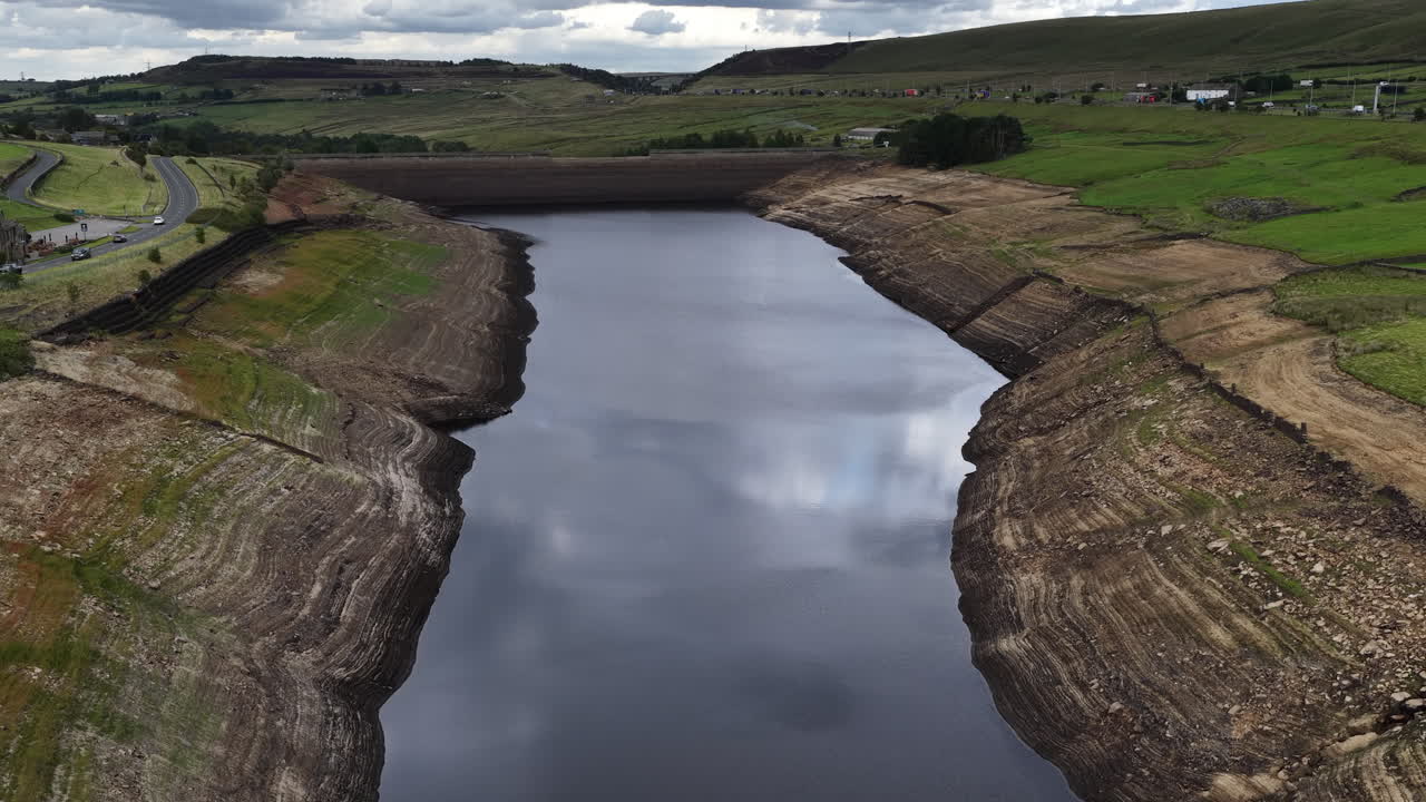 Aerial drone shot of Baitings Reservoir, West Yorkshire, showing low water levels, exposed shoreline, moorland hills, and cloudy sky reflections in a rural landscape