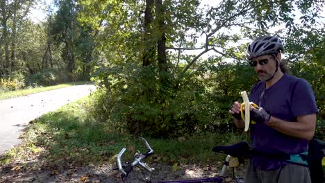 Mature-man-in-sunglasses,-helmet-and-gloves-next-to-his-bike,-eating-a-banana-on-the-side-of-the-road