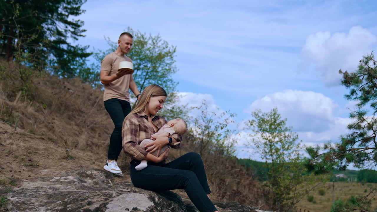 Blonde long-haired woman sits on the stone with baby in hands. Man comes up to them bringing a cake.