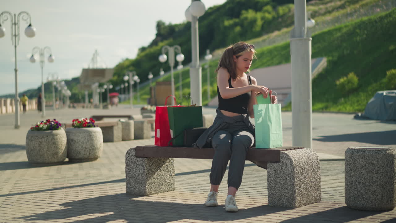 Lady seated on outdoor bench with shopping bags on both sides, retrieving a book from a mint green bag and placing a brown book back into the same bag, background includes lamp poles