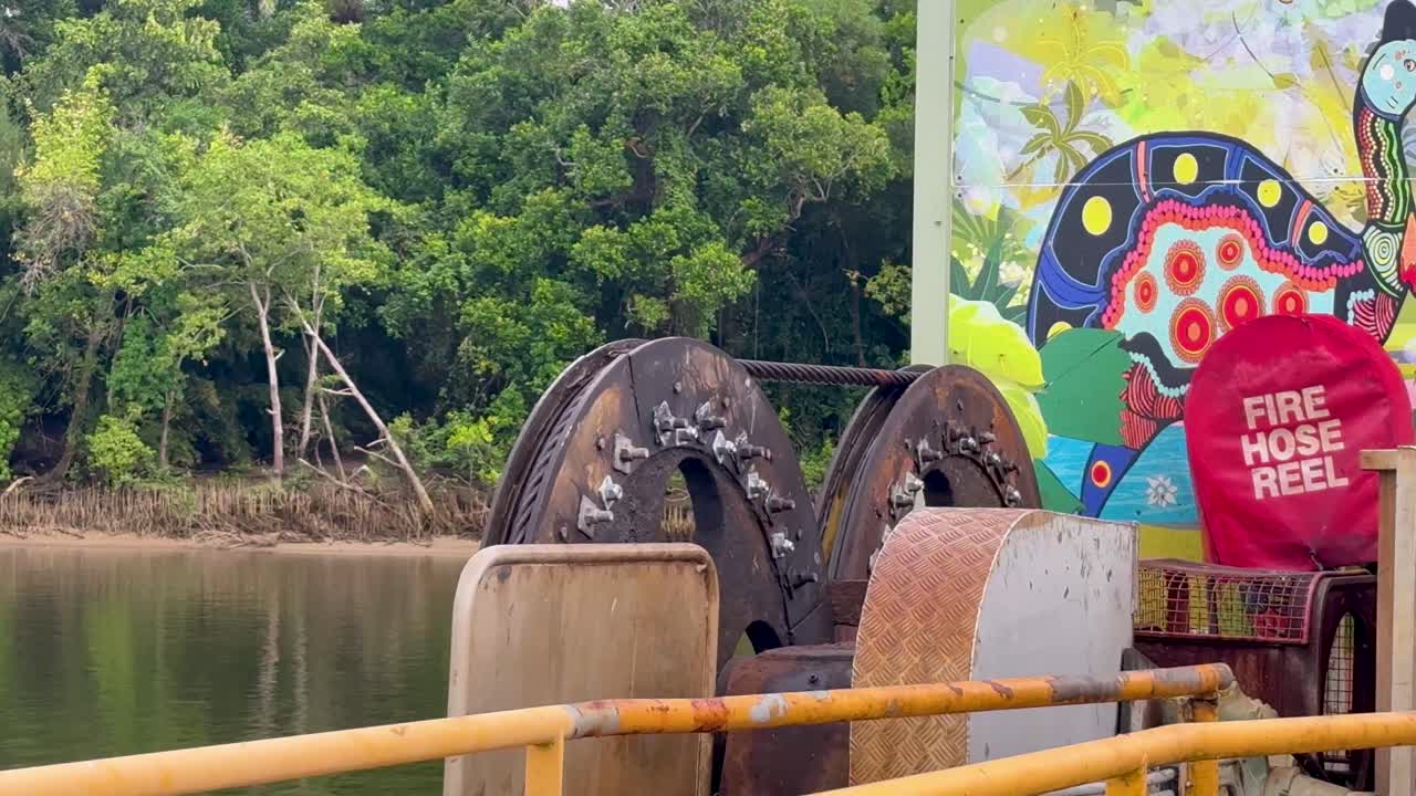 Ferry cable mechanism operates beside river, vibrant indigenous mural, lush tropical rainforest background, daylight