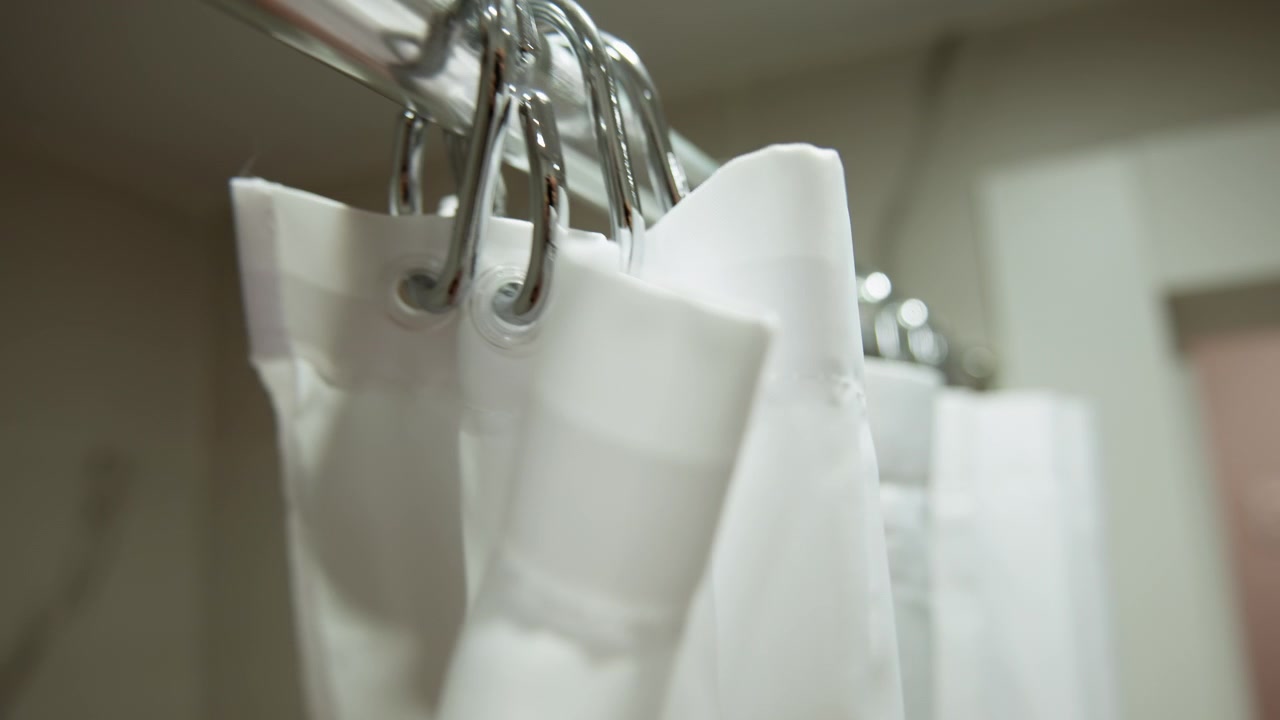 Primo piano di una tenda da doccia in tessuto bianco appesa ad anelli di metallo su un'asta da doccia curva, che mostra un leggero ondeggiamento e un delicato movimento verso la telecamera con pieghe diagonali, luce soffusa e un ambiente domestico tranquillo.