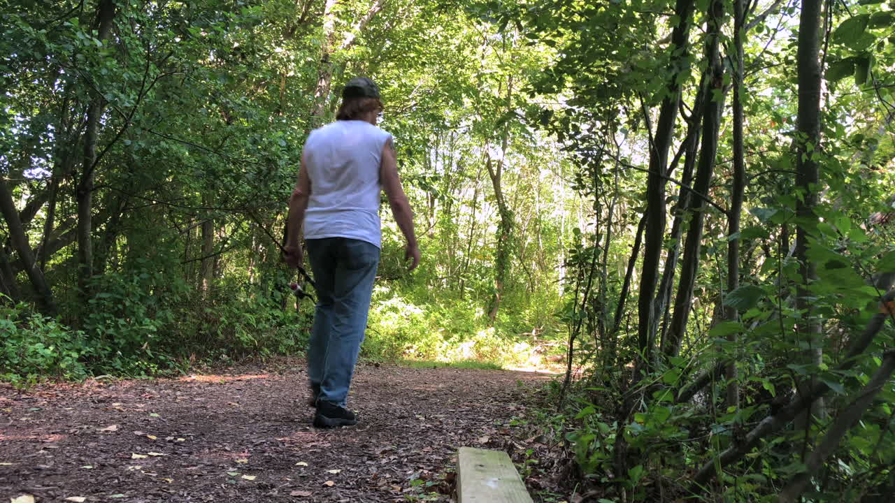 A man walking away from the camera down a path holding a fishing pole