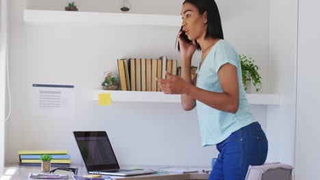 Mixed-race-gender-fluid-person-talking-on-smartphone-working-from-home-using-a-laptop