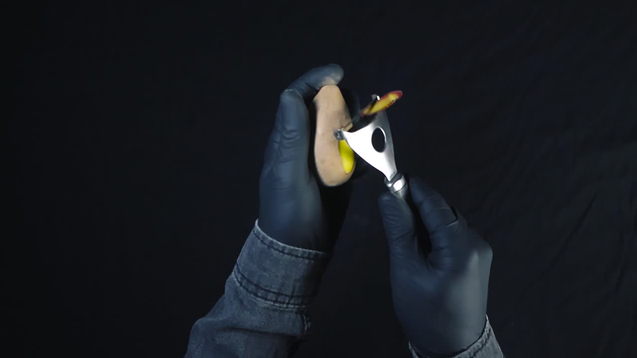 Top down view of a chef or a cook with nylon black gloved peeling a fresh and dirt covered potato in front of a black background. Potato peels falling in slow motion and golden yellow potato revealed