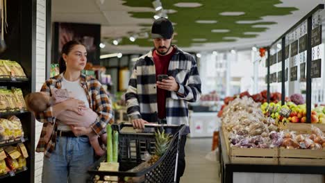 A-man-in-a-plaid-shirt-looks-at-the-grocery-list-on-his-phone-with-his-wife-and-small-infant-while-shopping-in-a-supermarket