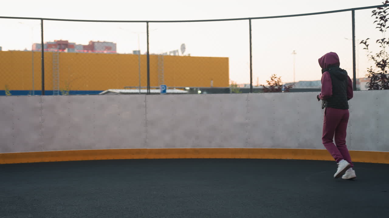 Hoodie wearing athlete jogging around circular sports court past white post against urban highrise background at sunset, pacing in padded vest and matching jogger pants with white sneakers on surface