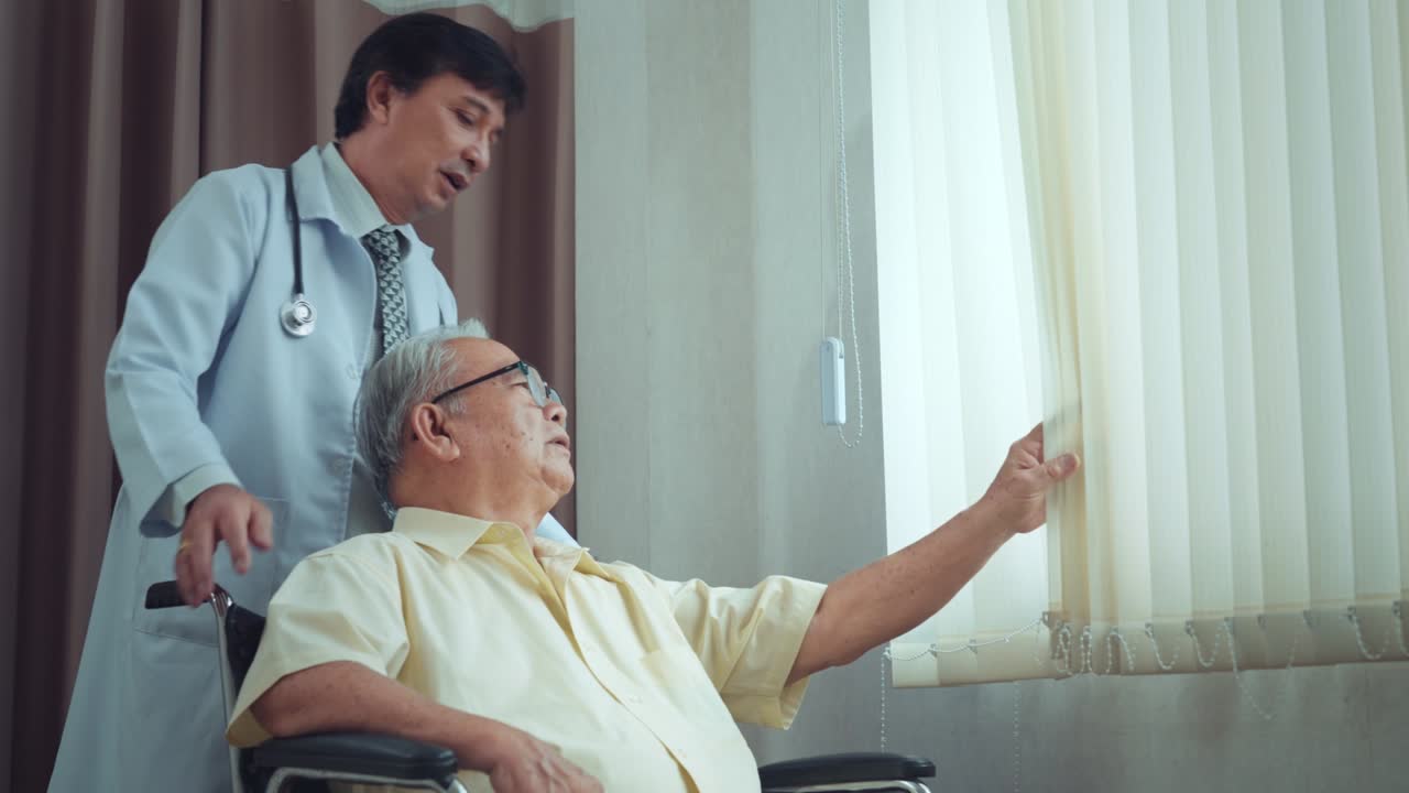 Male asian doctor encourages a patient grandfather in a wheelchair while looking out the window at nursing home in hospital. older people healthcare support concept, hope