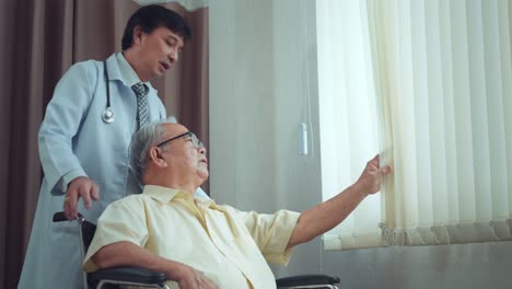 male asian doctor encourages a patient grandfather in a wheelchair while looking out the window at nursing home in hospital. older people healthcare support concept, hope