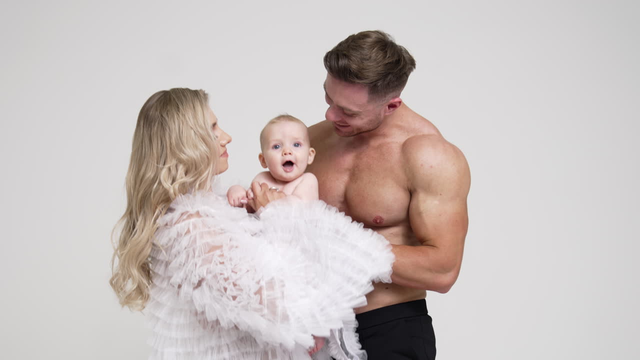 Caucasian family having little baby. Man with bare torso and blonde woman in white blouse with their son in studio. White backdrop.