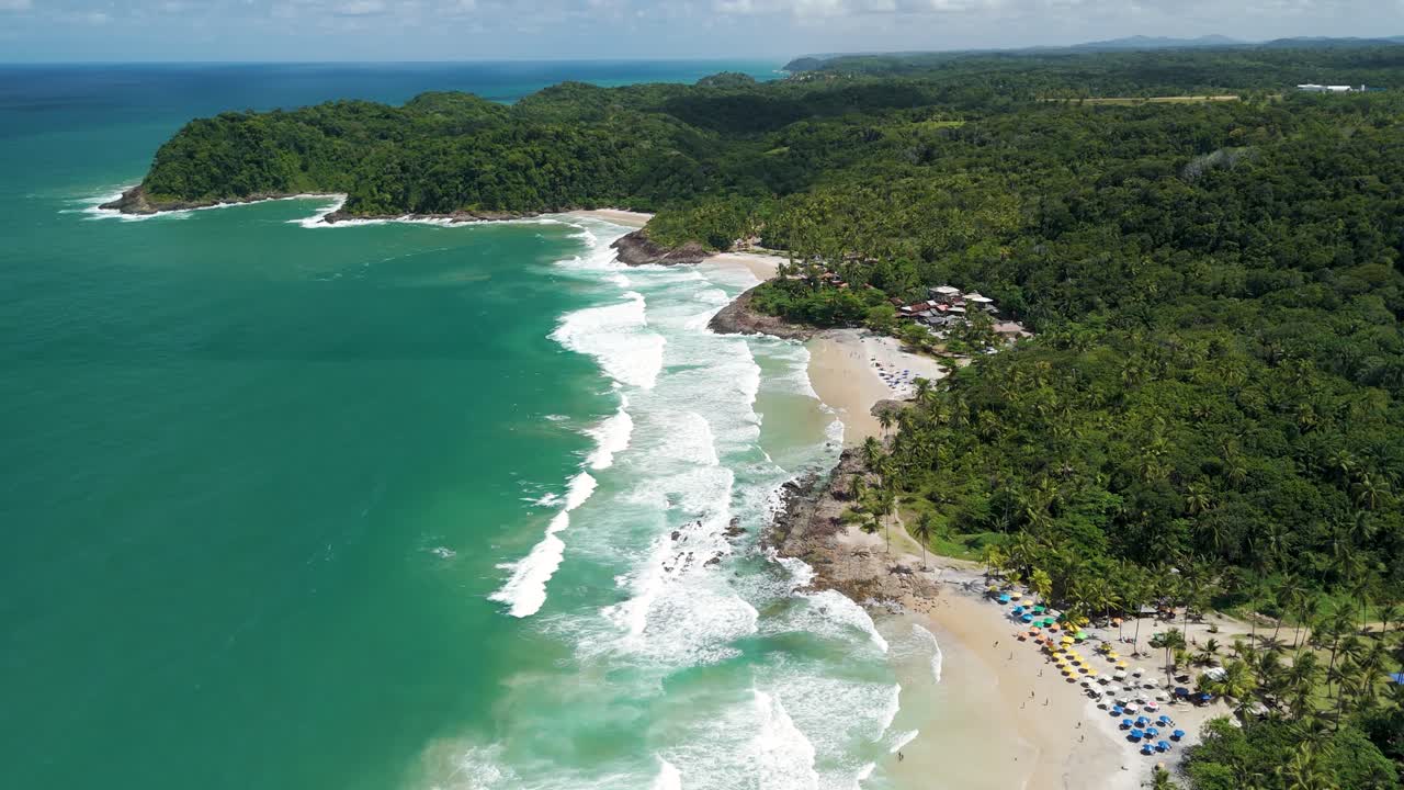 Aerial of turquoise waves lapping against tropical beaches with forest on sunny day, wide