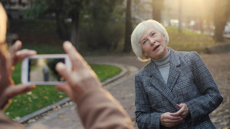 senior woman posing in the park while her husband taking a photo of her with smartphone camera