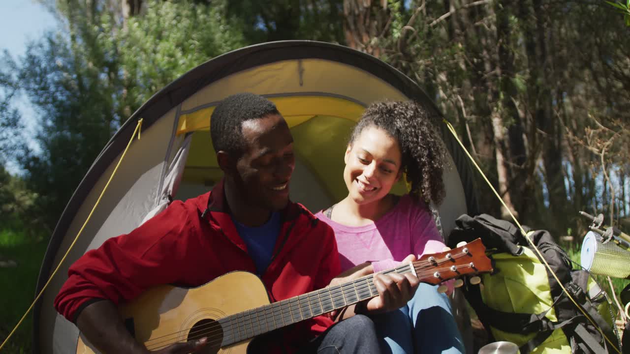 una pareja diversa sonriente sentada en una tienda de campaña y tocando la guitarra en el campo