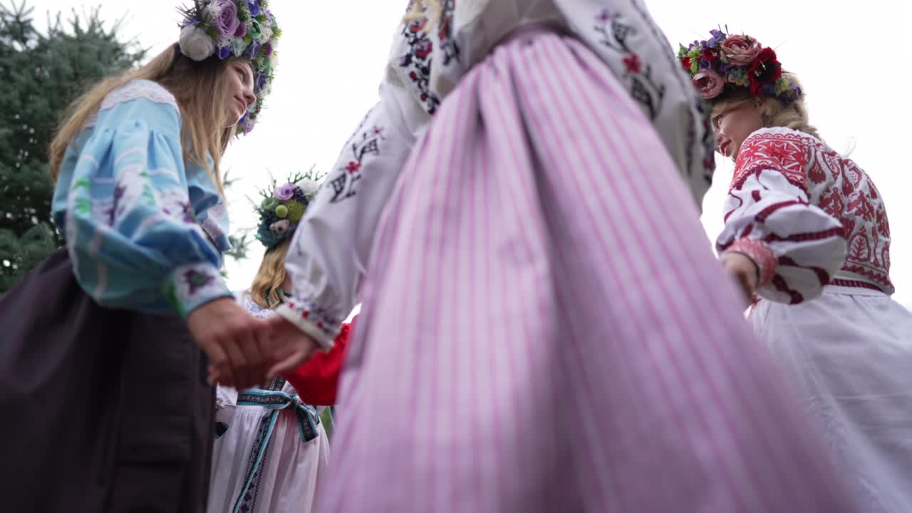 Bottom angle view of smiling adult woman standing outdoors as circle of young ladies in Ukrainian traditional dresses walking around holding hands. Happy lady enjoying folk dance in park. Slow motion.