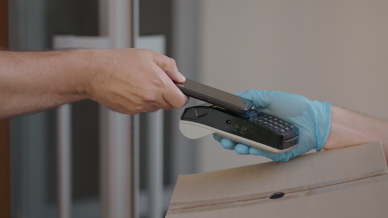 A male deliveryman in gloves brings a box and gives the terminal to pay for the purchase. Carry a box with an NFC credit card payment terminal or mobile phone. Contactless payment