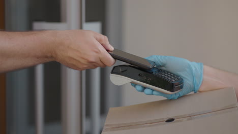 un repartidor masculino con guantes trae una caja y da la terminal para pagar la compra. llevar una caja con una terminal de pago de tarjeta de crédito nfc o teléfono móvil. pago sin contacto