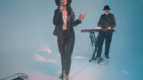 a vocalist in a black leather jacket, trousers, and hat stands at a vintage microphone held with one hand. a keyboardist plays a red sampler against a blue background as the camera zooms in and out