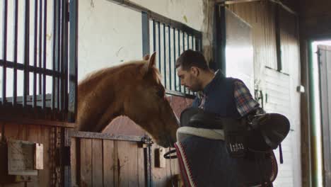 man is stroking a horse in stable while holding a saddle.