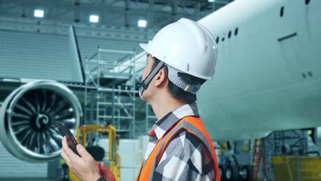 vista lateral cercana de un ingeniero masculino asiático con casco de seguridad usando un teléfono inteligente y mirando a su alrededor mientras está de pie con el avión en el hangar