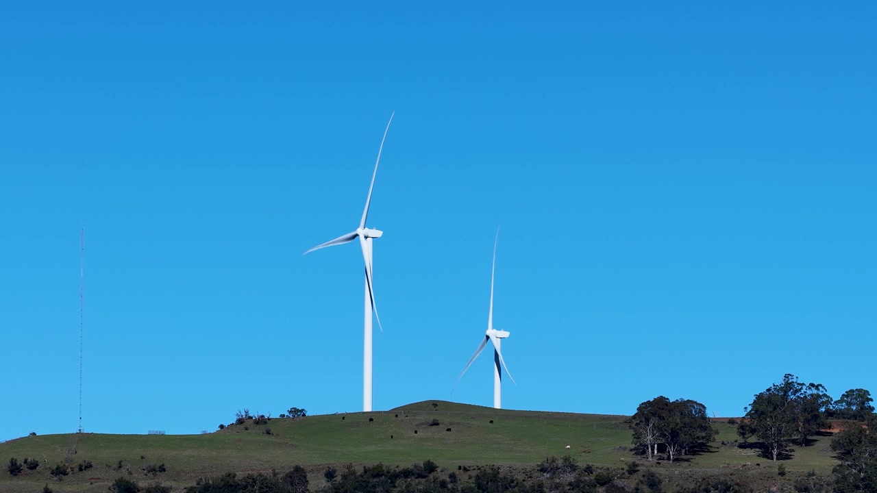 Inquadratura ampia statica di due turbine eoliche che generano energia rinnovabile su un crinale erboso.