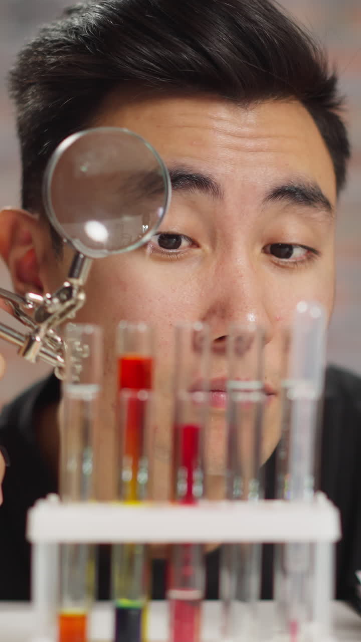 Curious Asian chemist man looks at test tubes with liquids through magnifying glass doing tests in scientific laboratory close view