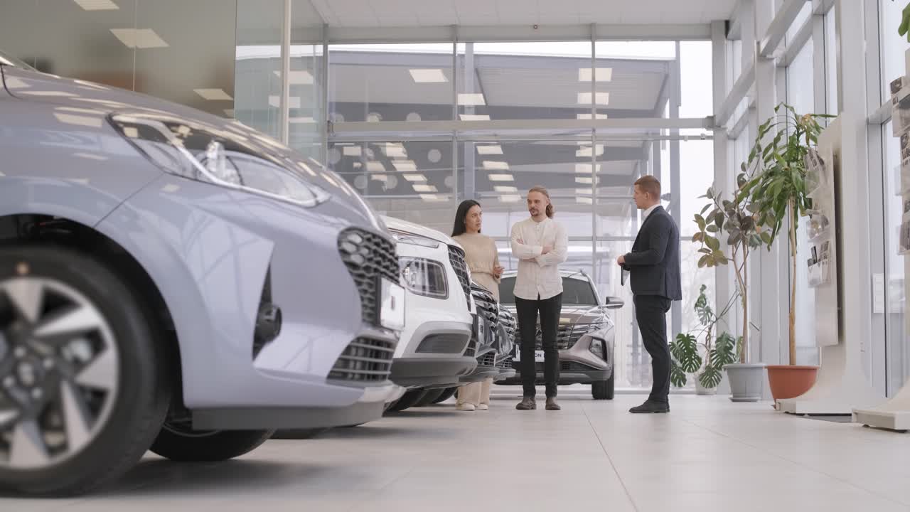 un joven vendedor de coches mostrando a una pareja de jóvenes un nuevo automóvil en el salón de concesionarios.