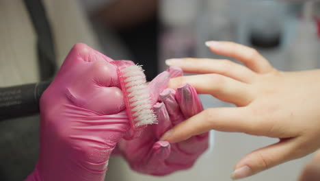 close up view of nail technician wearing pink gloves brushing and filing client's fingernails with precision using handheld tool and soft bristle brush during detailed manicure process