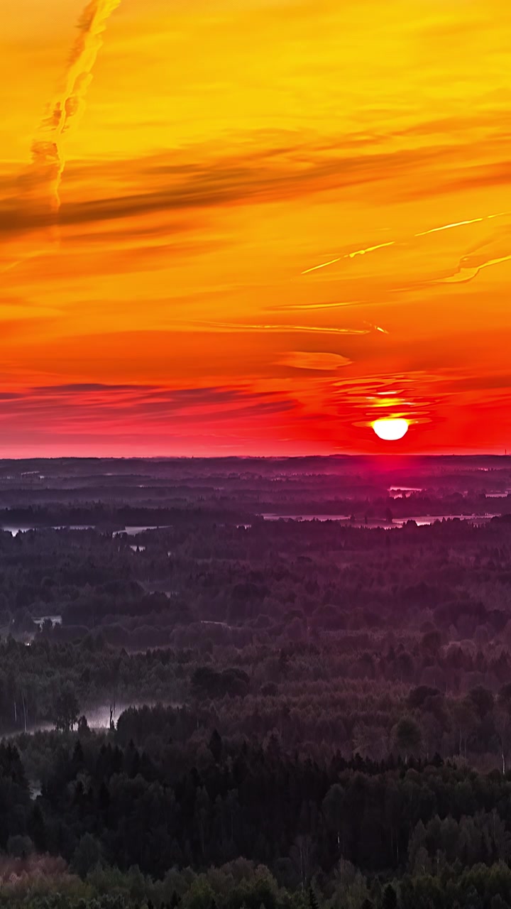 Vertikale Luft-Zeitrafferaufnahme eines feurig-orangen Sonnenaufgangs mit aufgehender Sonnenscheibe über dunklem, nebligem Wald, mit Sonnenstrahlenreflexion und Nebel in Tälern bei Sonnenaufgang am flachen, dunstigen Horizont.