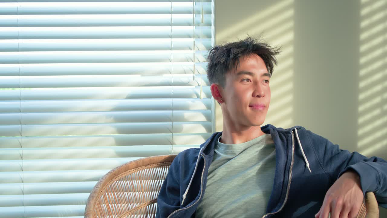 フード付きの服を着て、日差しが差し込むブラインドのそばの家で、籐の椅子に座って、振り返り笑顔を見せるアジア人男性。