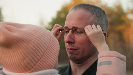 a touching outdoor moment between a father and daughter as she adjusts his glasses while embracing him. the man, wearing a green jacket, looks at her lovingly while she stands close