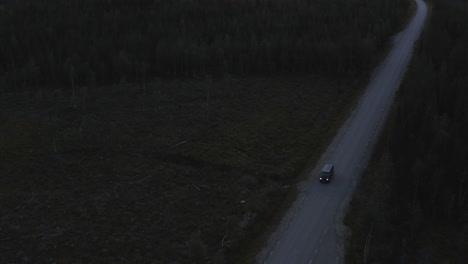 4k drone shot of a car driving on a country road at dusk through forest with headlights