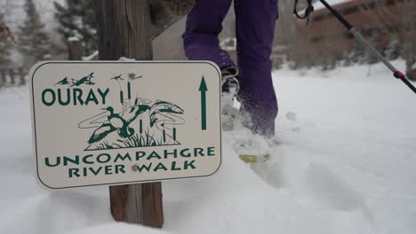 Close-up,-female-legs-in-showshoes-walking-by-Ouray-Uncompahgre-River-Walk-sign-in-snow-capped-landscape