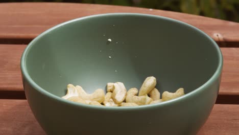 plant based ingredient cashew nuts falling into green bowl outside in slow motion on wooden picnic table - 4k food clip