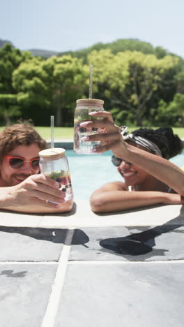 vertical video: diverse couple enjoying drinks by pool at home, greenery around