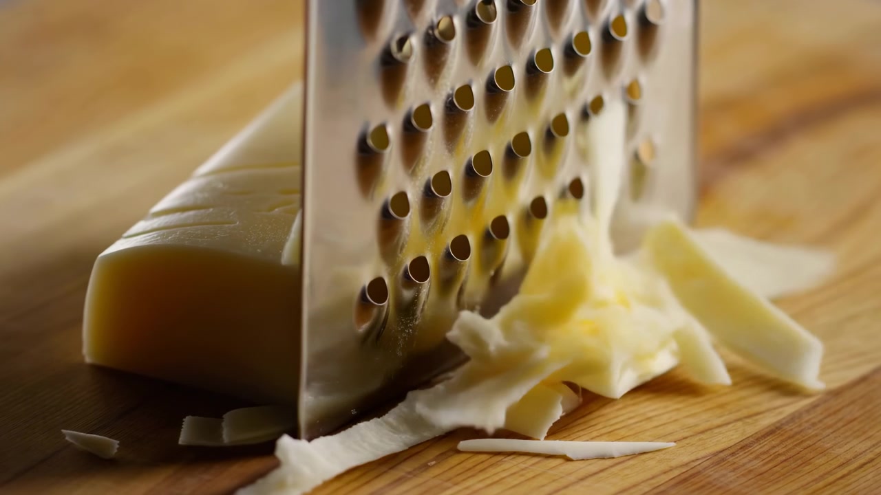 Tight close-up showing stainless box grater pressed against pale yellow semi-hard cheese on warm wooden board while fine shreds gather below, suggesting fresh cooking preparation, with cheese, grater