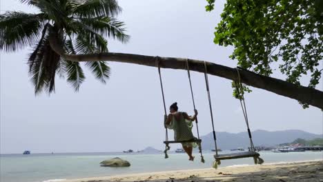 a woman swings leisurely on a beach swing hung from a palm tree in phuket, thailand. the serene scene features turquoise waters, sandy shore, and tropical foliage under soft daylight