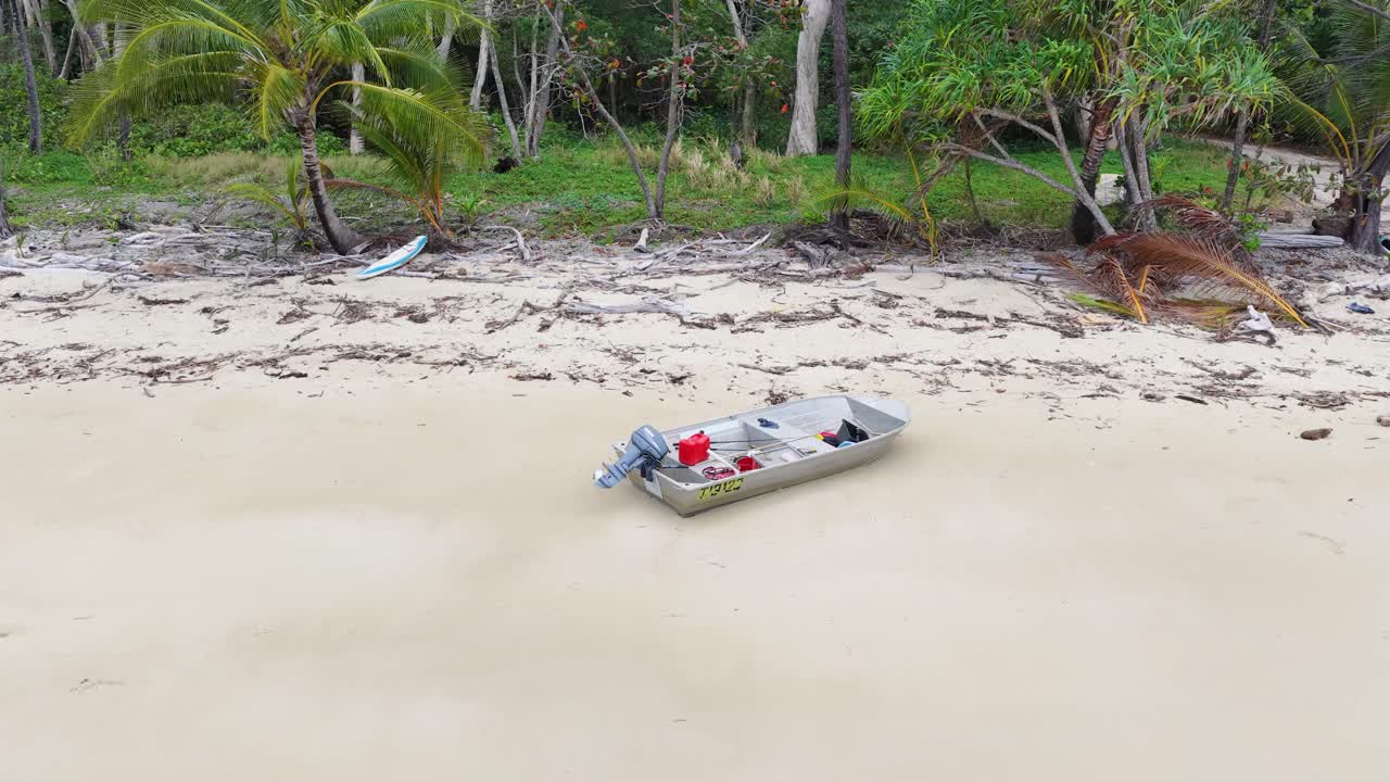 Drone flies over empty aluminum boat stranded on sandy beach, lush rainforest background, daylight