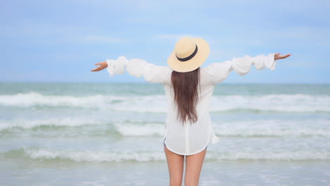 joyful woman with long hair and floppy hat raising and spreading hands in front of waves and horizon of tropical sea, back view, full frame, slow motion