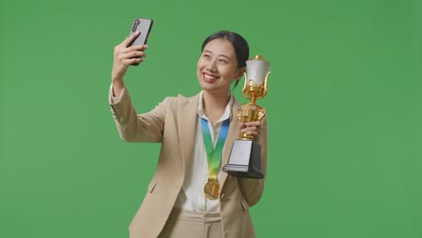 금메달을  슈트를 입은 아시아 사업가 여성이 금 트로피를 들고 스마트폰으로 사진을 찍으며 스튜디오의 녹색 화면 배경에서 첫 번째 우승자로